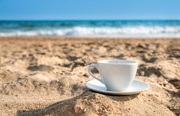 white cup with tea or coffee on sand beach front of sea, close up